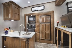 The Signature Ultra Lite 8297S displays the optional American Maple cabinetry and Chocolate fabric palette. Galley features include solid-surface countertops and an under-mount sink, along with a microwave oven, a gas oven, and a three-burner stove.